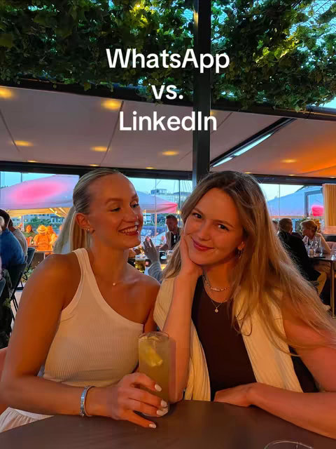 Two young women are sitting at a table outdoors, likely at a restaurant or bar. The main focus is on them, with a sign behind them. They are smiling and appear to be enjoying each other's company. The background features out-of-focus people, the sky, and greenery, suggesting an outdoor setting, likely during the day.