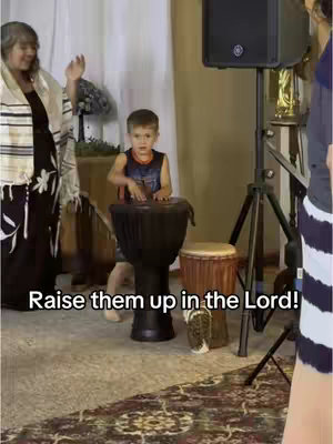 A young boy is drumming on a large drum in a room, with adults in the background and overlaid with the text 'Raise them up in the Lord!'.