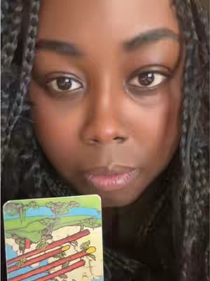 A close-up selfie featuring a Black woman with long, dark, braided hair. She looks directly at the camera with eyes that convey a slightly serious expression. She holds a tarot card in front of her, featuring colorful imagery of a landscape with red sticks. The background and her own features are softly illuminated.