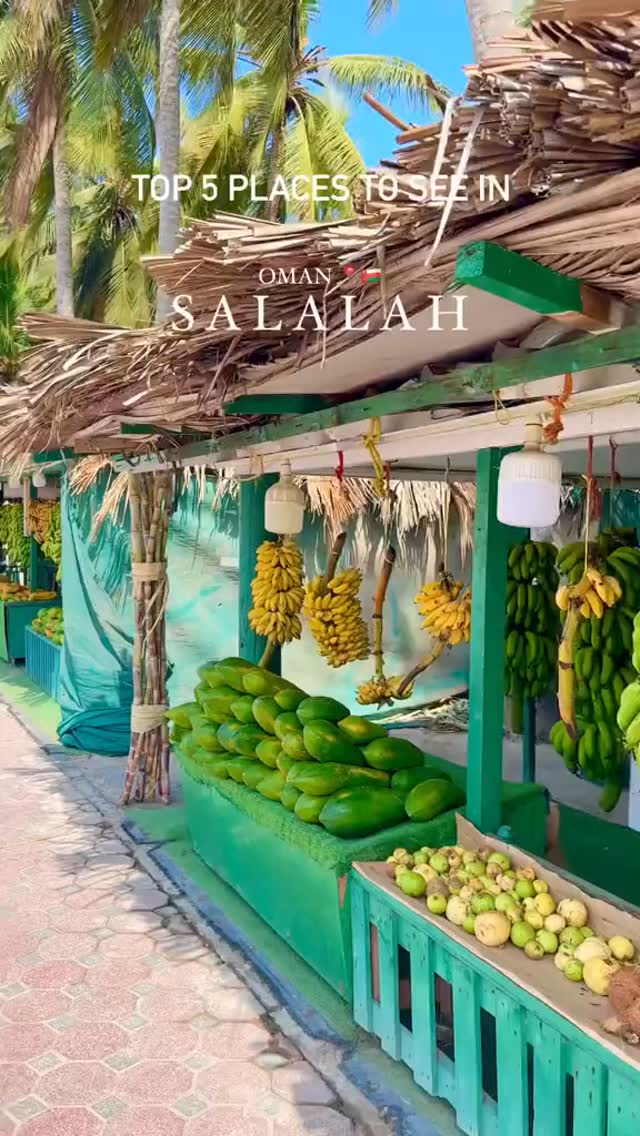 The image showcases a vibrant fruit stand in Salalah, Oman. The composition is dominated by fresh produce: green papayas are neatly stacked on a counter, while bunches of ripe bananas hang above. The stand is constructed with wooden beams and a thatched roof, giving a rustic feel. Lush palm fronds and a clear blue sky form the backdrop.