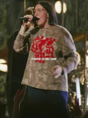A woman with glasses and short, dark hair sings into a microphone. Behind her, another person is slightly visible, and the background is softly lit.