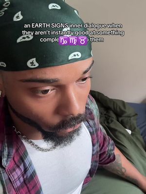 A close-up shot of a man with a beard, wearing a white shirt under a red and purple plaid shirt. He has a green bandana on his head, with text overlaid that says "An EARTH SIGNS inner dialogue when they aren't instantly good at something comple" followed by zodiac symbols. He appears to be looking down with a thoughtful expression.