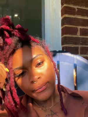 A close-up shot of a person's face with radiant skin and vibrant red locs and hair. The lighting is warm, with sunlight casting a glow. The face shows a calm expression. A gold necklace adorns the neck, and the background hints at an outside setting with a brick wall.