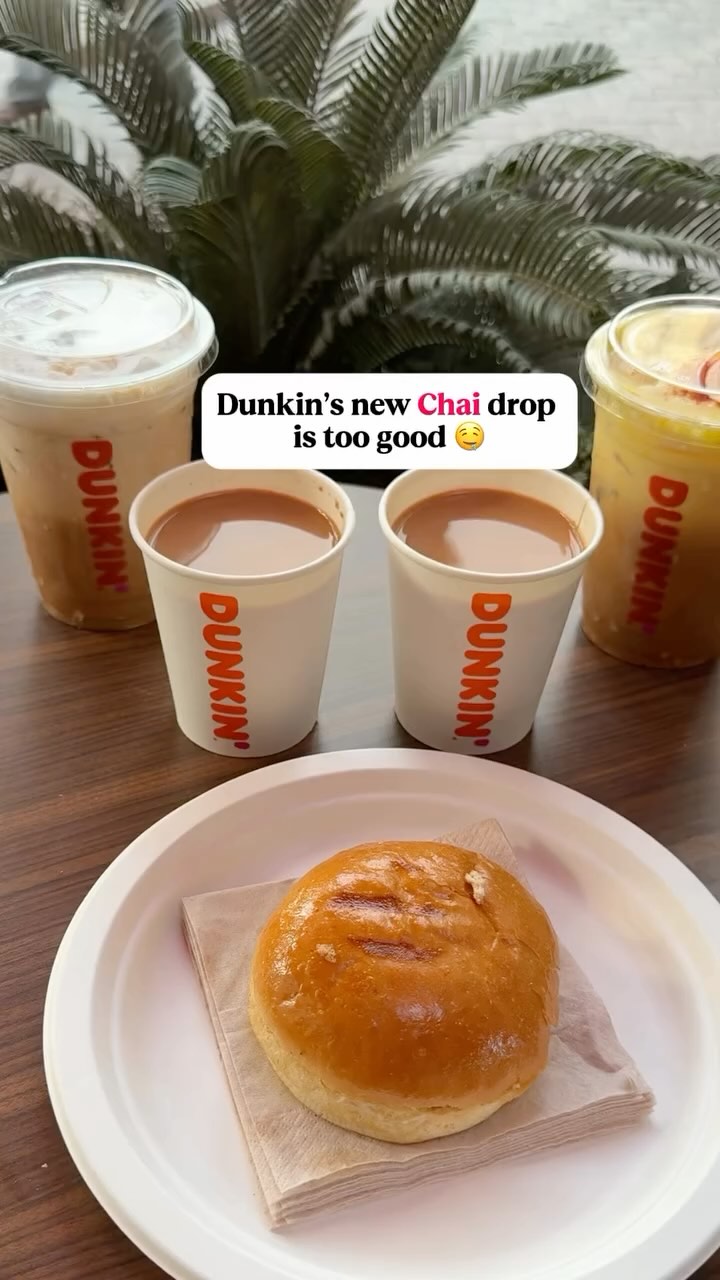 The image shows several Dunkin' beverages, hot and iced, alongside a plate with a golden-brown bun. Two cups hold hot chai, while other drinks are in clear cups, showing a pale beverage. The setting is on a wooden surface, with a leafy backdrop adding depth. The text overlay states, "Dunkin’s new Chai drop is too good".