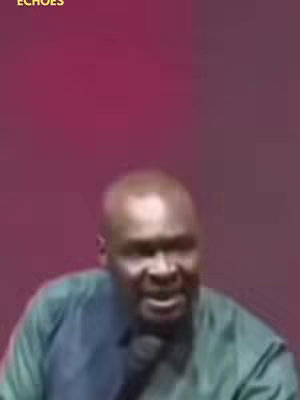 A man with a bald head speaking into a microphone is centered in the frame. The background is a solid, dark pink. The man is wearing a green shirt.