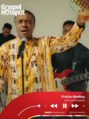 The cover image showcases a live performance with a male singer in center, wearing a yellow and brown patterned shirt. He's holding a microphone and singing with passion, his eyes closed. Behind him, two other men are playing instruments, including a guitar. The background is simple with a light-colored wall. A graphic at the bottom features a red playbar, song details and the Gospel Hotspot logo.
