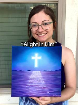 A woman with glasses smiles while holding a painting of a glowing cross above a body of water. Text "Alight In Him" is overlaid on the picture.