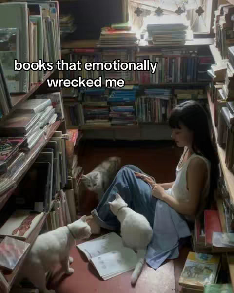 A young woman sits on the floor amidst bookshelves filled with books. She's surrounded by several cats, some of which appear to be interacting with an open book. The setting is dimly lit, giving a cozy and intimate feel, with a window providing some light.