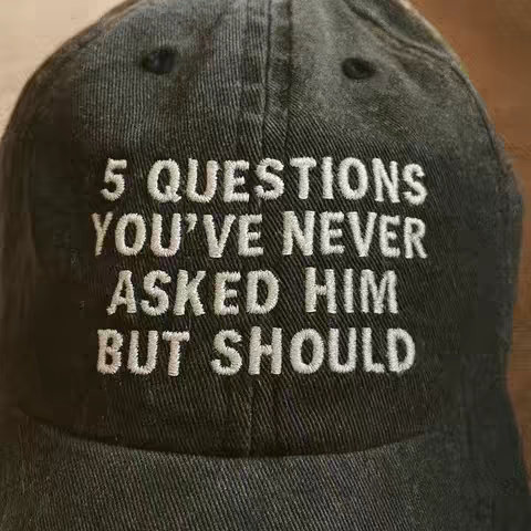 The image shows a close-up of a black baseball cap. The cap has white embroidered text across the front that says: "5 QUESTIONS YOU'VE NEVER ASKED HIM BUT SHOULD". The cap appears to be made of a textured material. The focus is sharp on the text, and the overall image has a soft, natural lighting.
