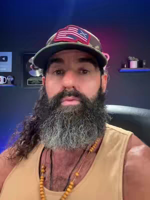 A man with a full beard is centered in a selfie-style shot. He's wearing a camo baseball cap with an American flag emblem, a tan tank top, and several beaded necklaces. He's in a room with dark blue walls, where a couple of plaques are visible on the left. The background is somewhat blurred.