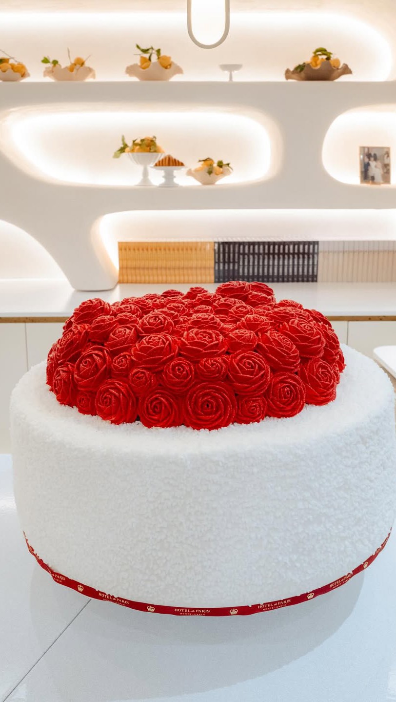 The image is a close-up of a rose cake in a boutique setting. A large cake sits on a white surface, the top entirely covered in red roses. The cake sits on a wide, cylindrical base covered in white, fluffy material. The base is trimmed with a red ribbon with gold text. The background includes white shelves holding pastries, and some other light colored decorative elements. The lighting is bright and even, highlighting the detailed textures.