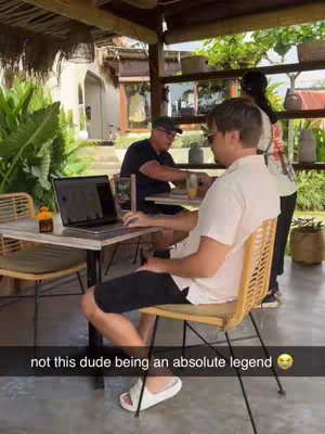 A man is seated at a table, working on a laptop located outdoors in what appears to be a tropical setting. He is wearing sunglasses, a light-colored shirt, and black shorts. Behind him, two other people are partially visible. The setting includes a table, chairs, and foliage. The composition is casual and candid, presumably taken in a relaxed environment.