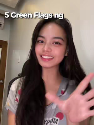 A young woman with dark, long hair is smiling at the camera, holding her hand up with her fingers open. Her face is the central focus, but elements of a room, including what appear to be an air conditioning unit, are visible behind her, the background is blurred. Text overlayed on the image says, '5 Green Flags ng'.