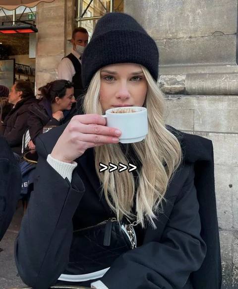 A woman with blonde hair wearing a black beanie and a black jacket is sitting outdoors, holding a white coffee cup and looking at the camera. The background includes a cafe setting with other people blurred out.