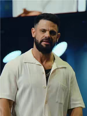 The image is a medium shot of a man, likely in his 40s, against a softly lit stage background. He is wearing a cream-colored, short-sleeve button-down shirt over a black undershirt, and a gold chain is visible. The man's complexion is warm-toned, and he has a close-cropped beard and hair. He seems to be speaking, with a slight expression suggestive of a sermon or speech in front of a crowd. The lighting is soft and diffused, with two large, white oval stage lights positioned behind the man, adding a gentle glow.