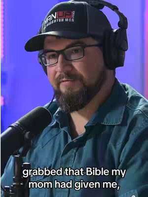 A man with a beard, glasses, and a cap sits in front of a microphone. He has headphones on and is looking towards the camera. The background is a blurry blue color.