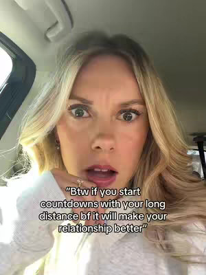 A woman is inside a car. The woman has pale skin and light-blonde wavy hair framing her face. Her open mouth shows her upper teeth slightly. She wears a white sweater and a silver ring with a small stone or embellishment. The background is blurred and shows the interior of a car and a seatbelt. The woman is looking at the camera.