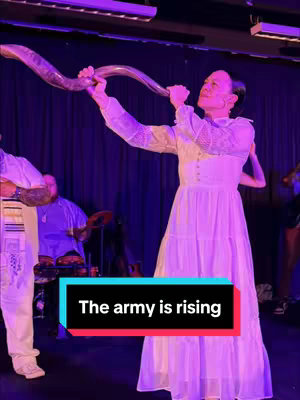 A woman in a white dress is blowing a shofar, a traditional Jewish horn. She is in front of a dark purple backdrop, with others visible in the background, including a person playing drums. The image has a vertical layout, with a hook at the bottom that reads 'The army is rising'. The lighting is dim with purple and blue hues.