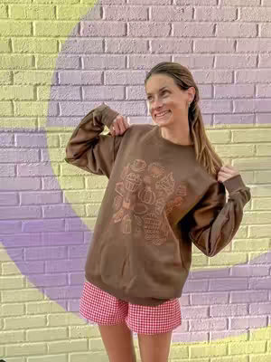 A woman stands in front of a brick wall painted with a yellow semi-circle and a purple semi-circle. She wears a brown sweatshirt with a cream-colored graphic on the front. The graphic looks like a bouquet with pumpkins, coffee cups, and other fall-themed items. She is also wearing red and white checkered shorts. She looks happy and is holding her sweatshirt.