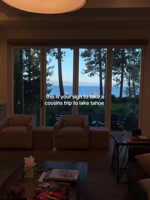 The image displays a living room with a large glass door showing a scenic view of a lake and trees. The room is furnished with couches, armchairs, and a coffee table. A text overlay suggests taking a trip to Lake Tahoe with cousins.