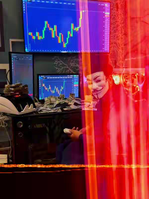 The image showcases a person wearing a Guy Fawkes mask, sitting in front of multiple computer screens displaying financial charts. Money is scattered on the desk, and the scene is partially obscured by vertical red light trails. The overall impression is one of digital trading or financial activity.