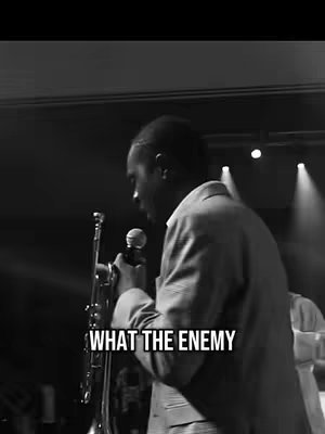 A grayscale image of a man in a suit jacket, holding a microphone in one hand and a trumpet in the other. He is on stage, with spotlights and other figures in the background. Text overlay at the bottom reads, 'WHAT THE ENEMY'.
