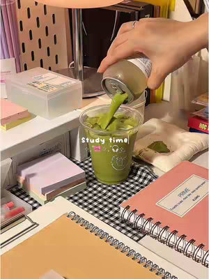 The cover image depicts a well-lit desk setup with various study supplies. A hand pours green matcha from a can into a clear plastic cup containing ice and matcha, with the text overlayed on the cup reading "Study time". The desk has a clear container, pink sticky notes, a notebook, and a pen with a lid. The background includes a checkered pattern and various study accessories.