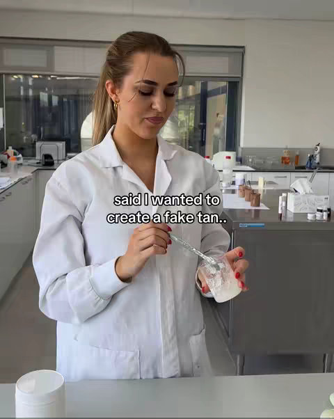 A woman in a white lab coat is standing in a well-lit laboratory setting. She is holding a clear beaker with a white substance in it, and a small metal spoon. The text overlay reads, "said I wanted to create a fake tan..". The background shows stainless steel counters and equipment.