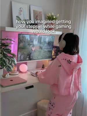 A young woman in a pink hoodie and sweatpants is seated at a gaming setup. She is facing away from the camera, interacting with a computer screen that displays a game. The setting includes a desk, a monitor, a keyboard, and decorative items, all illuminated by soft lighting. The overall aesthetic is clean and modern, with a focus on pink tones.
