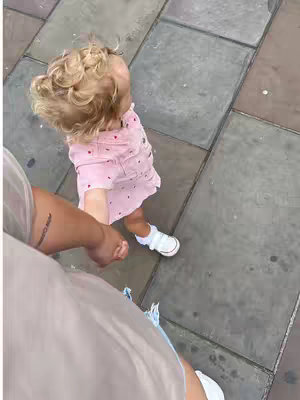 An overhead shot of a child walking on a gray stone surface, holding hands with another person who is partially visible on the left side of the frame. The child, facing forward with curly blonde hair, wears a pink dress covered in small red hearts and white shoes. The person holding the child's hand has a visible tattoo on their arm. The ground is made of large, rectangular gray tiles.