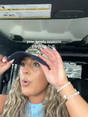 A woman inside a car, looking upward with a thoughtful expression, is the main subject. She is wearing a baseball cap and a light-colored shirt. The overhead view suggests a driving scene.