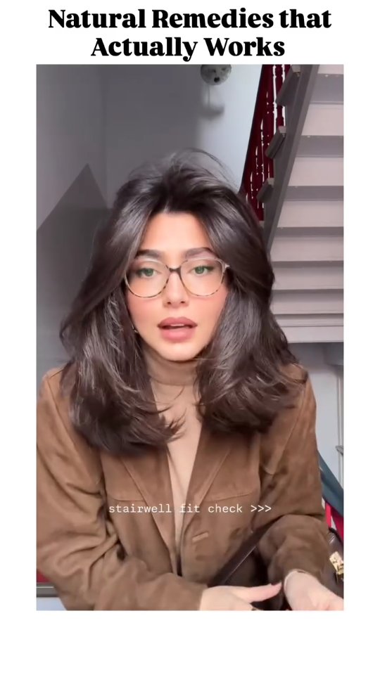 The image features a woman with long, dark hair, wearing glasses and a brown jacket, standing in front of a staircase. Text overlays read "Natural Remedies that Actually Works" and "stairwell fit check >>>". The composition is well-lit, with a neutral background.