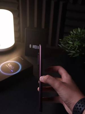 A close-up shot of a violet smartphone held in a person's hand, showcasing its side profile with the power button visible. The hand, with a smartwatch, holds the phone against a dark background with a blurred box and other objects. A bright white lamp illuminates the scene, creating a contrast.