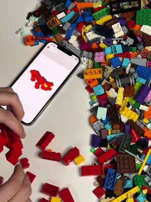 A top-down view shows a smartphone with a Lego dragon model on the screen, surrounded by a scattered array of colorful Lego bricks on a white surface.