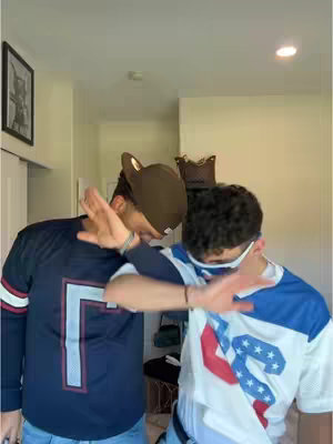 Two men, likely boyfriends, strike a pose indoors. They are wearing matching arm gestures. The man on the left is wearing a navy blue jersey with a white 'L' on the chest, and a brown cap. The man on the right is wearing a white and blue jersey with a red and white star on the chest. Overall, the image is well-lit.