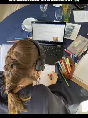 A person, viewed from behind, is seated at a table with a laptop and papers, studying with headphones on. The table is covered with items including books and pens. The colors are muted, with the focus on the central figure and the items related to studying.