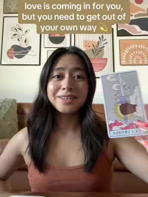 A woman with dark hair and a red crop top holds up a tarot card. Text overlayed on the image reads, 'love is coming in for you, but you need to get out of your own way.' Various framed pictures hang behind her.
