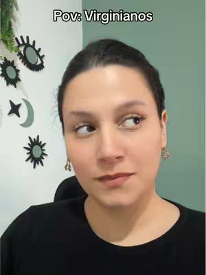 A woman with dark hair gazes to the side with a thoughtful expression. She is wearing a black top, and gold earrings. Behind her, there's a wall with a combination of decorative shapes, including a crescent moon and stars.