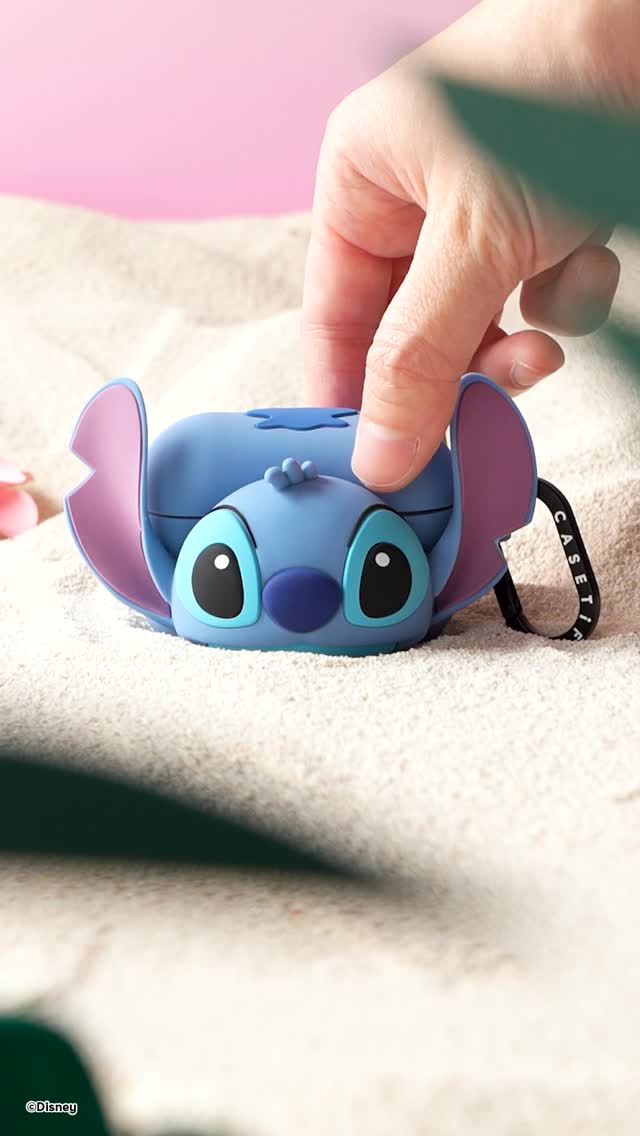 The image features a blue Stitch-themed AirPods case resting on white sand. A hand is positioned above the case, as if about to open it. The background includes a faded pink and sky-blue gradient. The overall composition conveys a playful, tropical theme, with the focus on a collectible product placement.