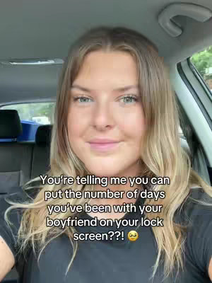 A woman is shown in the front seat of a car, gazing directly at the camera with a gentle smile. The background hints at an outdoor setting with a glimpse of a tree, suggesting daytime. The overall image is well-lit, with natural light enhancing her facial features.