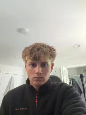 A selfie-style shot featuring a young man with blond hair and a black jacket, set against a neutral background. The man's head and shoulders fill most of the frame, with the upper part of the image showing the ceiling and lights. The composition is simple, with soft lighting and a focus on the subject's face, which has a neutral expression.