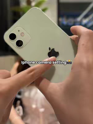 The image shows a person holding a light green iPhone, angled towards the viewer. The phone's camera lenses and the Apple logo are visible. The person's hands are in the foreground, and the background is blurred. A text overlay, 'iPhone camera setting' with a camera emoji, is added at the bottom.