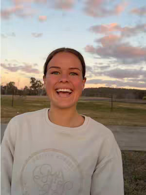 A young woman with fair skin and short, brown hair is centered in the frame, smiling widely with teeth visible. Her eyes are bright. She wears a light-colored sweatshirt. The background features a landscape with a field, a few trees, and streaks of color from the sunset.
