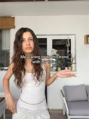A young woman with long, wavy brown hair stands in front of a window and white wall. The woman wears a white tank top, a decorative chain belt, and white pants. She has a neutral expression, with her arms extended outwards, as the text overlay indicates she is waiting to do a trend with her twin.