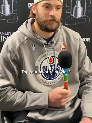 A man wearing an Edmonton Oilers hoodie and a white hat is holding a microphone with three color stripes. Text on the image asks "Top four favorite movies?". The background appears to be a press event setting.