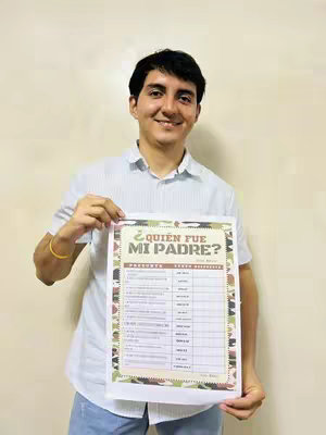 A young man with dark hair is holding a printed sheet with text on it. He is smiling broadly at the camera.The man is wearing a light blue button-down shirt. He is standing in front of a light tan wall.