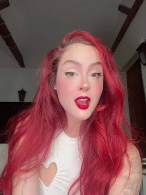 A woman with bright red hair and makeup looks towards the camera with an open mouth and red lipstick. She wears a white top with a heart cutout at the chest. The background is a dimly lit interior.