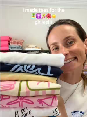 A smiling woman holds a tall stack of folded t-shirts in front of her. The shirts are various colors and patterns. The background shows a well lit room with shelves and additional clothing items.