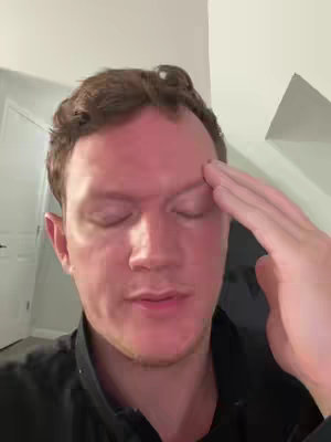 A man with short brown hair is seen close up, with his eyes closed and his hand raised to his forehead as if in distress or contemplation. The background shows a muted interior setting.