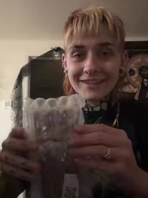 A person of ambiguous gender is smiling while holding a clear plastic container. The setting appears to be indoors, possibly in a room. The person is wearing multiple piercings and a choker. The background is blurred, but hints at an interior space with furniture.